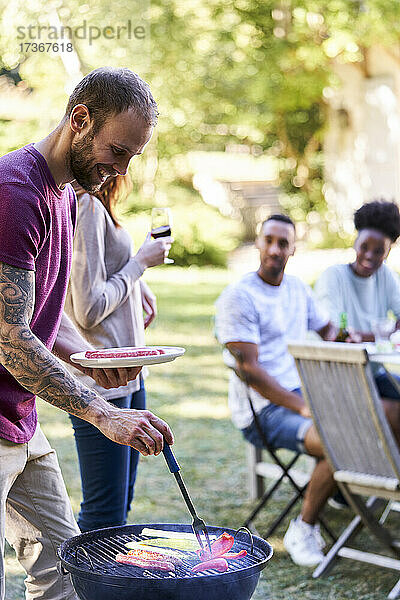 Junger Mann bereitet Essen auf einem Grill zu  während seine Freunde im Hintergrund etwas trinken