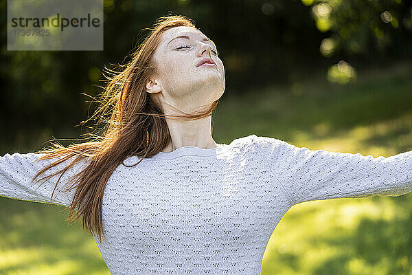 Junge Frau mit geschlossenen Augen streckt ihre Arme im Park aus