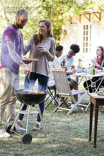 Junger Mann bereitet Essen am Grill zu  während seine Freunde am Tisch im Hintergrund sitzen