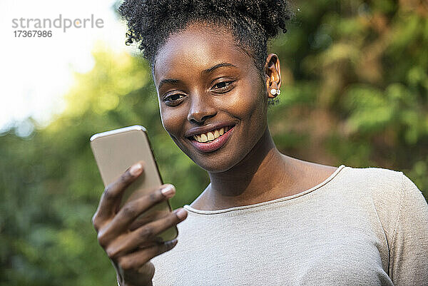 Nahaufnahme einer lächelnden jungen Frau  die im Park ihr Smartphone benutzt