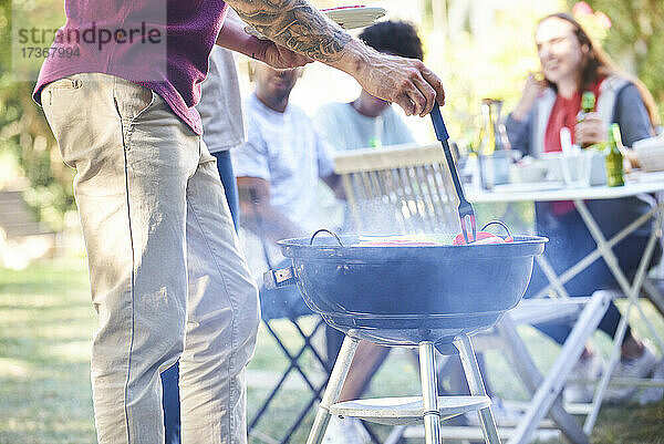 Junger Mann bereitet Essen im Grill zu  während seine Freunde im Hintergrund sitzen