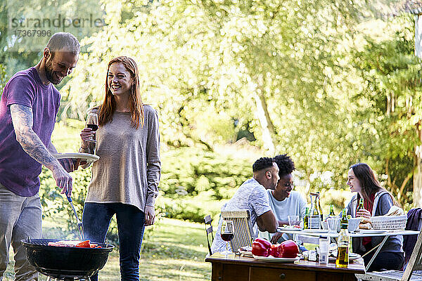 Junger Mann bereitet Essen auf einem Grill zu  während seine Freunde im Hintergrund etwas trinken