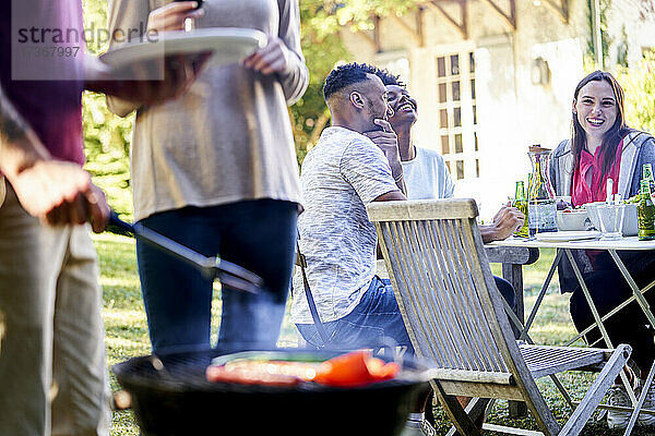 Junger Mann bereitet Essen am Grill zu  während seine Freunde am Tisch im Hintergrund sitzen