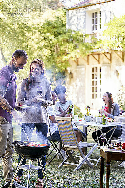 Junger Mann bei der Zubereitung von Speisen am Grill  während seine Freunde im Hintergrund Getränke zu sich nehmen