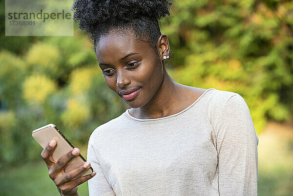 Nahaufnahme einer lächelnden jungen Frau  die im Park ihr Smartphone benutzt