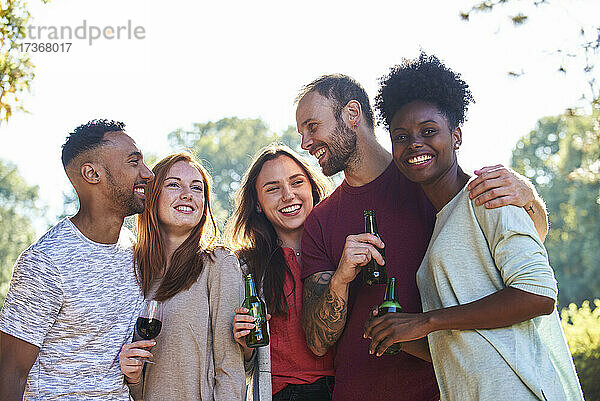 Lächelnde junge Freunde stehen mit Bierflaschen und Weinglas im Garten