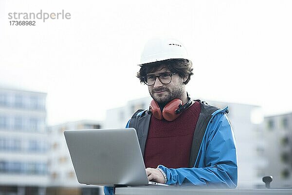 Junger Ingenieur mit Helm und Hörschutz checkt mit Laptop Arbeitstelle außen  Freiburg  Baden-Württemberg  Deutschland  Europa