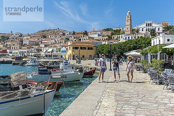 Drei Touristen  Fischerboote im Hafen von Chalki mit türkisblauem Wasser  Promenade mit bunten Häusern des Ortes Chalki  Chalki  Dodekanes  Griechenland  Europa