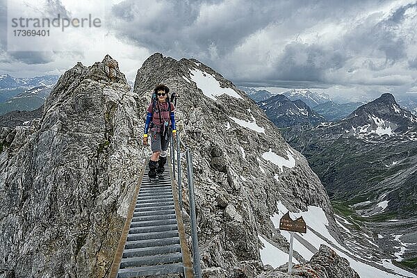 Wanderin auf einer Metallbrücke am Fels  Gratweg mit Schneeresten  Bergpanorama mit Gipfel Hohes Licht  dramatischer Wolkenhimmel  Heilbronner Weg  Allgäuer Alpen  Allgäu  Bayern  Deutschland  Europa