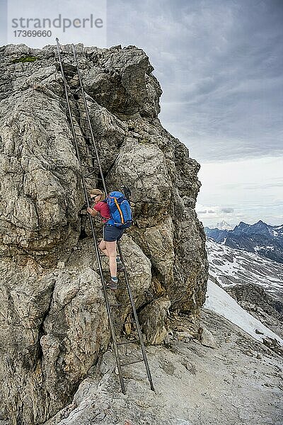 Wanderin besteigt Metalleiter am Fels  Bergsteiger auf Gratweg mit Schneeresten  dramatischer Wolkenhimmel  Heilbronner Weg  Allgäuer Alpen  Allgäu  Bayern  Deutschland  Europa