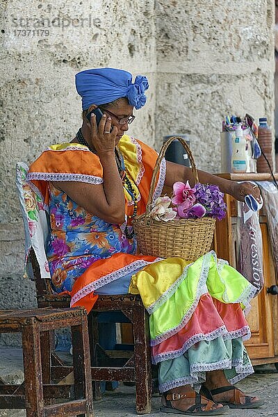 Frau  Kubanerin  in vielfarbiger Tracht  sitzt auf Stuhl und telefoniert mit Mobiltelefon  Altstadt  Hauptstadt Havanna  Provinz Havanna  Große Antillen  Karibik  Kuba  Mittelamerika