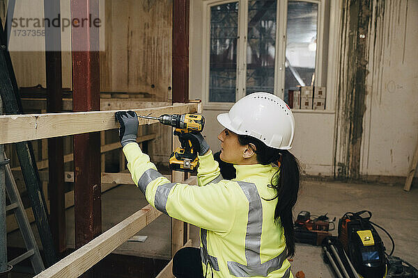 Weiblicher Arbeiter mit Schutzhelm bei der Arbeit auf einer Baustelle mit einer Bohrmaschine auf einem Holzbrett