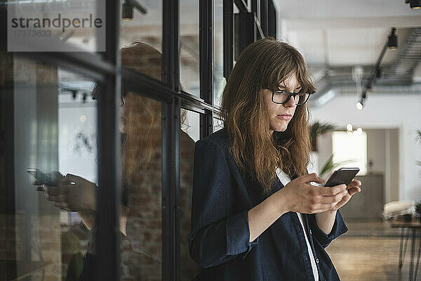 Junge Unternehmerin bei der Textnachrichtenübermittlung auf einem Smartphone in einem Start-up-Unternehmen