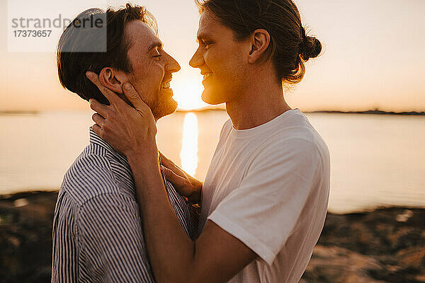 Lächelndes schwules Paar  das bei Sonnenuntergang am Seeufer von Angesicht zu Angesicht steht