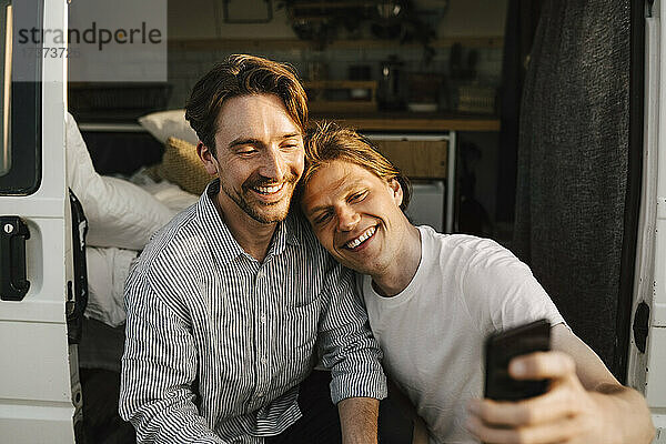 Lächelnde männliche Freunde machen ein Selfie mit dem Handy vor der Tür eines Campingwagens