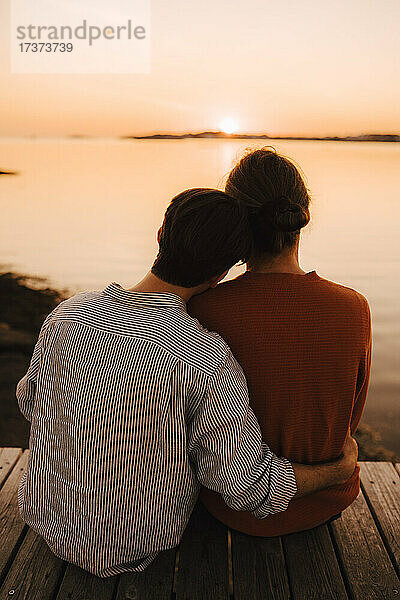 Rückansicht von männlichen Freunden  die den Sonnenuntergang betrachten  während sie auf dem Pier sitzen