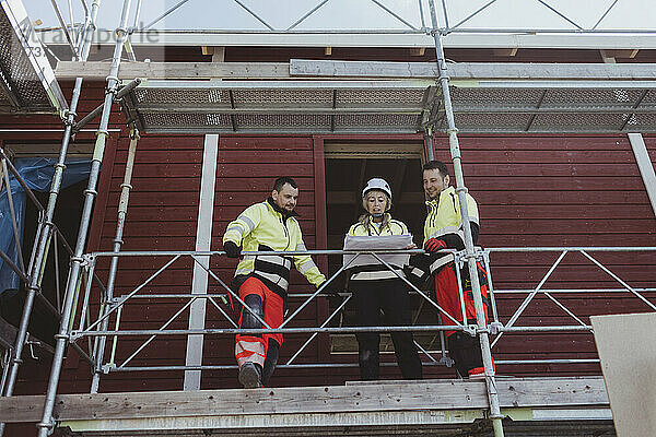Weibliche Bauunternehmerin diskutiert mit männlichen Kollegen auf einem Baugerüst über einen Grundriss