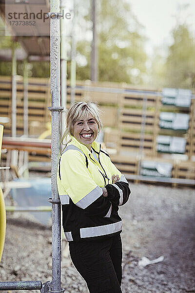 Porträt einer glücklichen Bauunternehmerin  die mit verschränkten Armen auf einer Baustelle steht