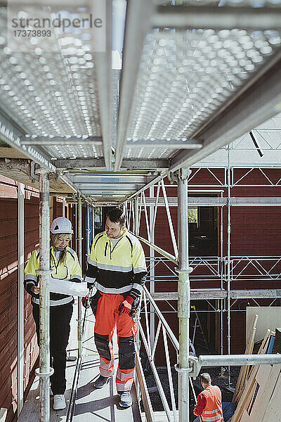 Bauunternehmerin in voller Länge  die mit einem männlichen Kollegen einen Grundriss bespricht  während sie auf einem Baugerüst läuft