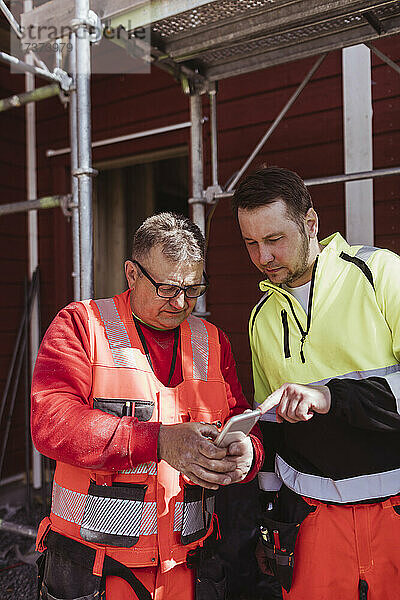 Älterer männlicher Bauarbeiter  der mit einem Kollegen auf der Baustelle über sein Smartphone diskutiert