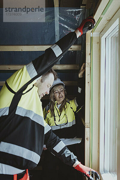 Eine Bauarbeiterin betrachtet einen männlichen Kollegen  der auf der Baustelle einen Fensterrahmen ausmisst