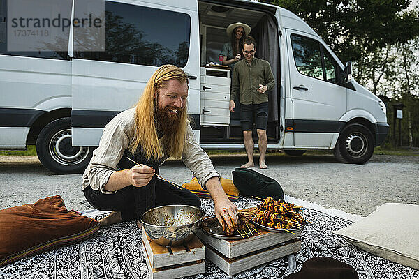 Bärtiger Mann bereitet Fleischspieße zum Grillen vor  während Freunde im Wohnmobil sitzen