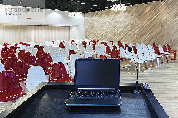Ein Laptop auf einem Podium und mehrere Stuhlreihen in einem großen Raum