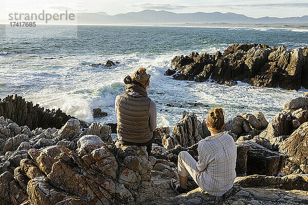 Frau und junges Mädchen sitzen am felsigen Ufer und blicken auf das Meer.