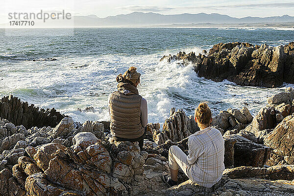 Frau und junges Mädchen sitzen am felsigen Ufer und blicken auf das Meer.