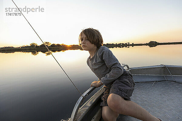Ein Junge angelt von einem Boot aus auf dem flachen  ruhigen Wasser des Okavango-Deltas bei Sonnenuntergang