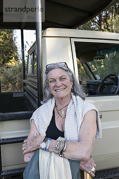 Ältere Frau steht lächelnd neben einem Safari-Jeep in Botswana.