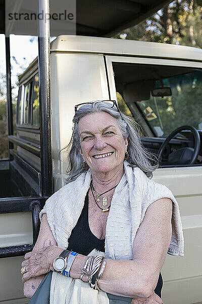 Ältere Frau steht lächelnd neben einem Safari-Jeep in Botswana.