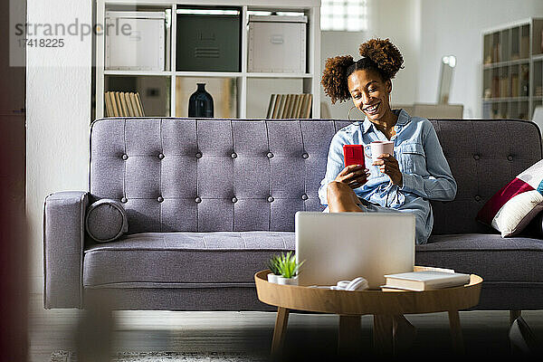 Junge Frau führt Videoanruf über Smartphone  während sie im Wohnzimmer Kaffee trinkt