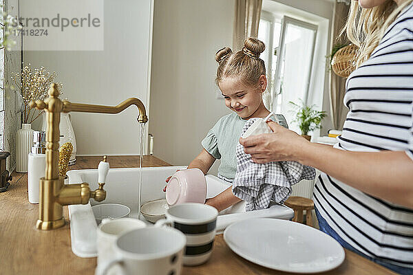 Lächelnde Tochter wäscht mit Mutter zu Hause Geschirr im Waschbecken