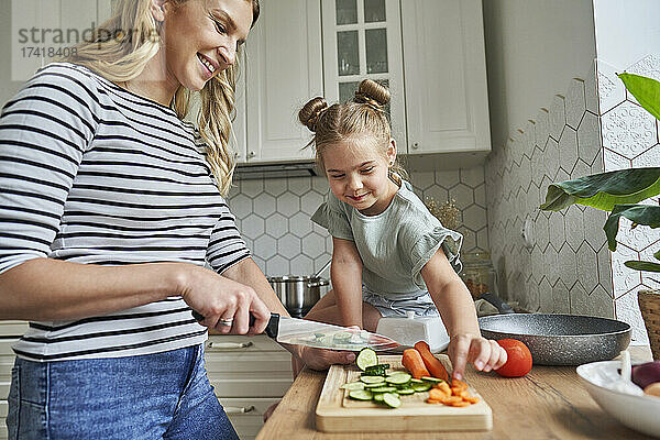 Lächelnde Mutter schneidet Gemüse  während ihre Tochter auf der Küchentheke sitzt