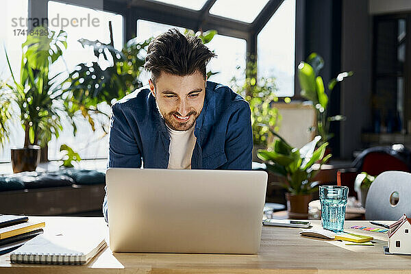 Lächelnder männlicher Profi  der einen Laptop benutzt  während er sich im Büro auf den Schreibtisch lehnt