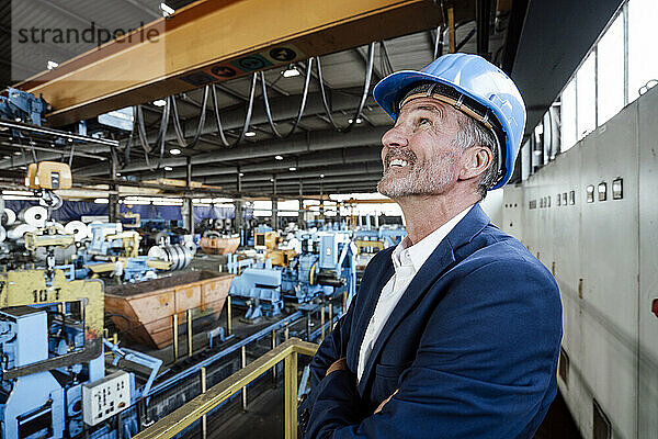 Lächelnder männlicher Ingenieur mit Bauarbeiterhelm  der auf den Laufkran im Stahlwerk blickt