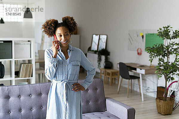 Lächelnde Frau mit der Hand auf der Hüfte  die im Wohnzimmer über ihr Smartphone spricht