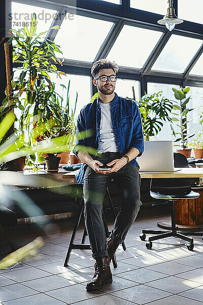 Männlicher Berufstätiger hält Smartphone in der Hand  während er am Schreibtisch im Büro sitzt