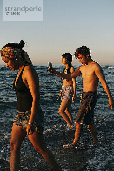 Männliche und weibliche Freunde gehen bei Sonnenuntergang durch das Meerwasser