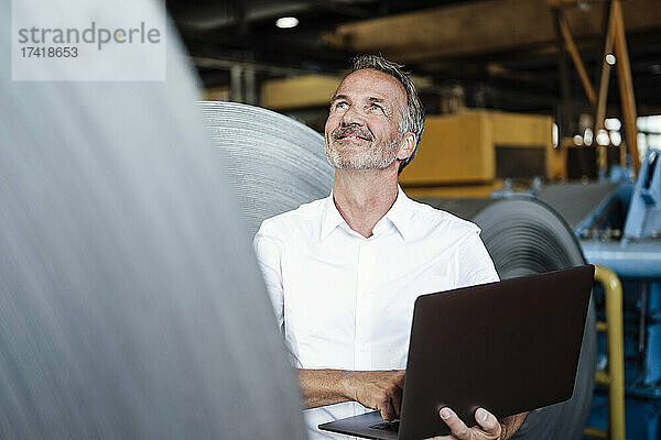 Lächelnder männlicher Inspektor mit Laptop  der die Blechrolle im Stahlwerk überprüft