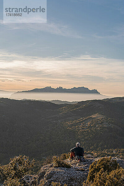 Älterer Mann blickt auf den Berg  während er beim Wandern an einer Klippe sitzt