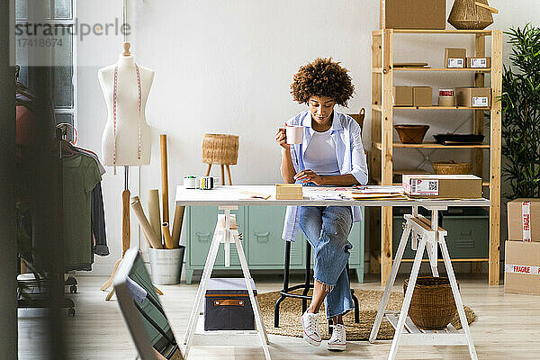 Modedesignerin mit Tasse sitzt am Schreibtisch im Studio