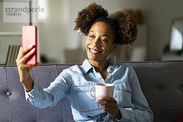 Lächelnde Frau  die zu Hause ein Selfie mit einer Kaffeetasse über ihr Mobiltelefon macht