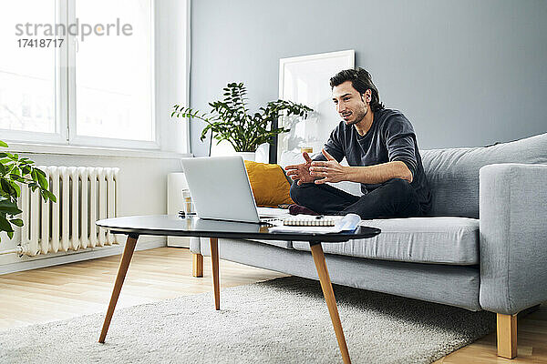 Männlicher Profi während eines Videoanrufs auf dem Laptop  während er zu Hause auf dem Sofa sitzt