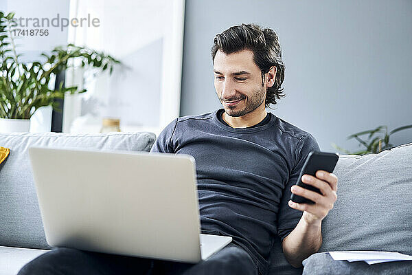 Geschäftsmann arbeitet am Laptop und hält sein Smartphone in der Hand  während er zu Hause sitzt