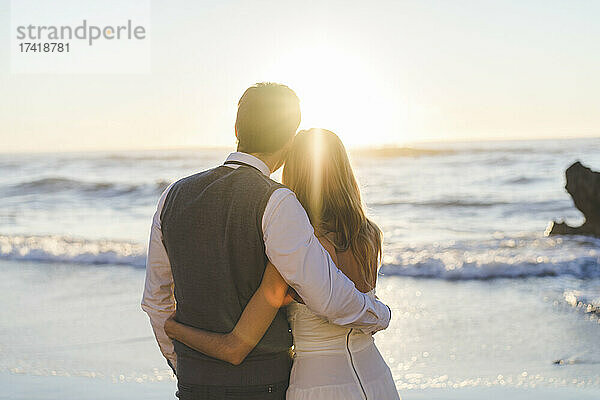 Junges frisch vermähltes Paar mit Arm um den Blick auf den Sonnenaufgang am Strand