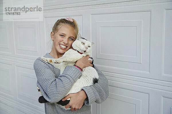 Blondes Mädchen umarmt Katze  während es an einer weißen Wand steht