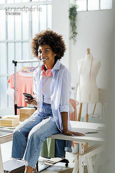 Lächelnde Geschäftsfrau mit Smartphone sitzt am Schreibtisch im Studio