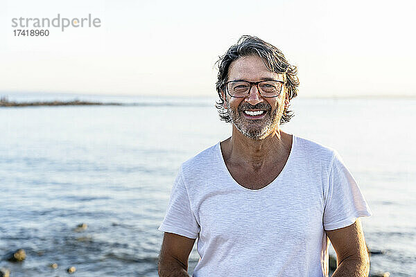 Glücklicher reifer Mann mit Brille am Strand bei Sonnenuntergang